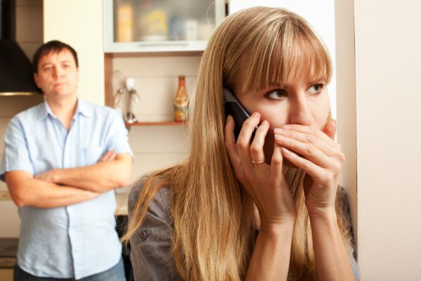Donna preoccupata al telefono copre con una mano la bocca, mentre un uomo osserva in piedi sullo sfondo.